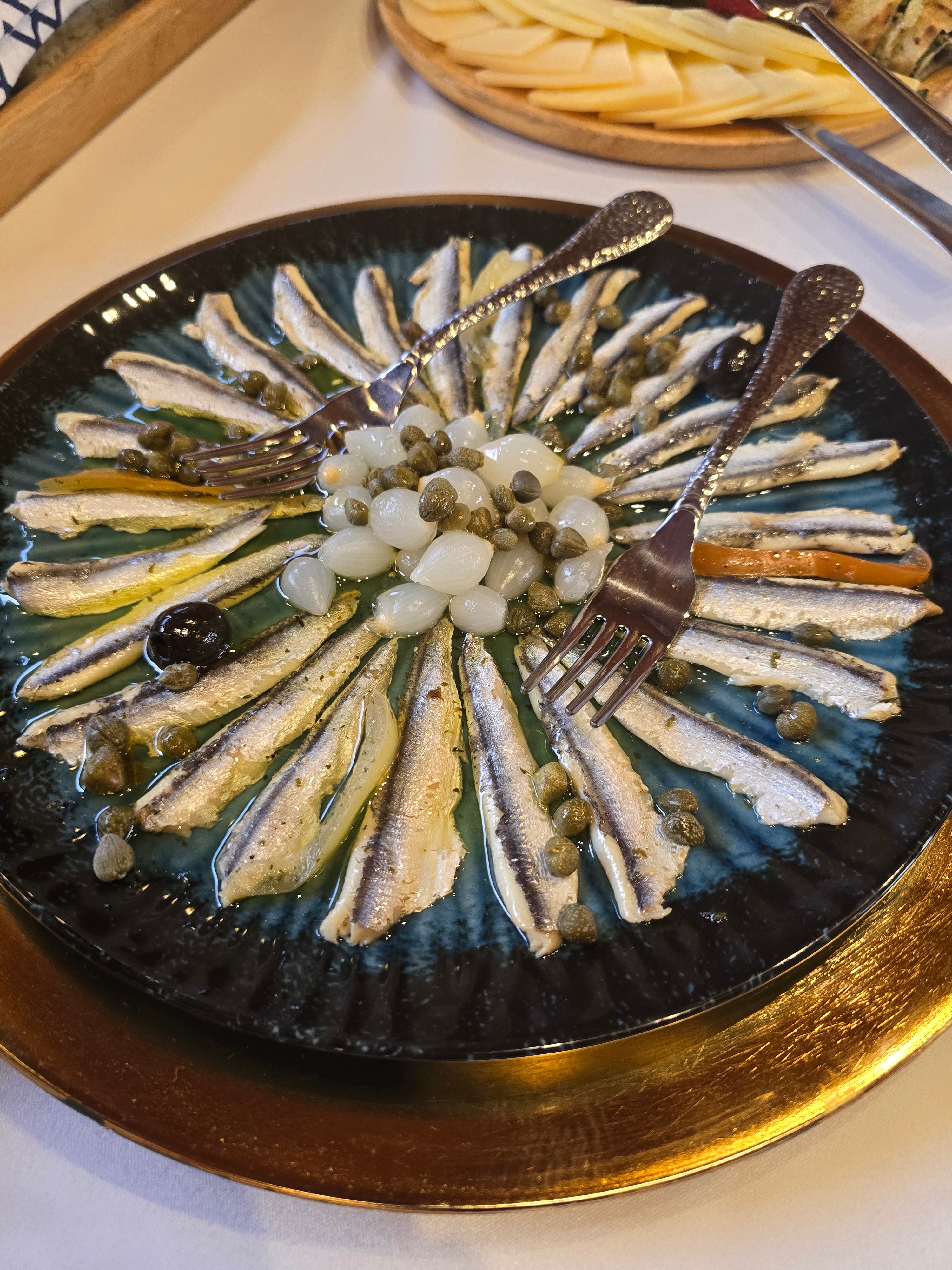 Marinated Adriatic anchovies arranged in a starburst with capers, pearl onions and olives on a dark ceramic plate