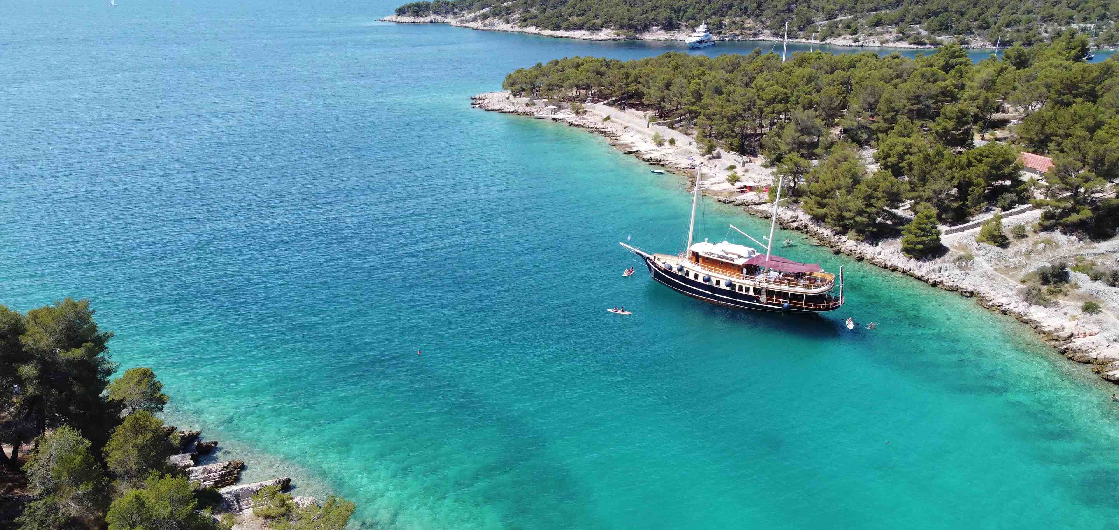 Aerial view of the yacht Polaris anchored in a secluded turquoise Adriatic cove ringed by pine-covered shores, with paddleboarders in the clear water beside her