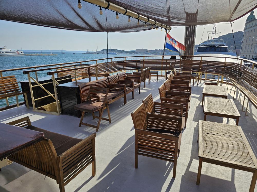 The open sun deck of Polaris with teak chairs, benches and tables under a shade canopy, overlooking Split harbor