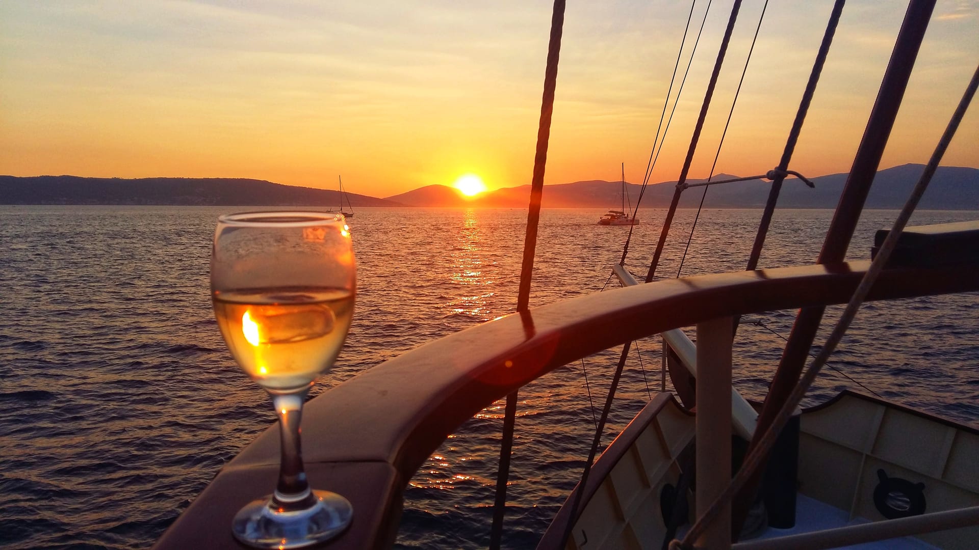 A glass of white wine on the rail with the sun setting over islands and sailboats